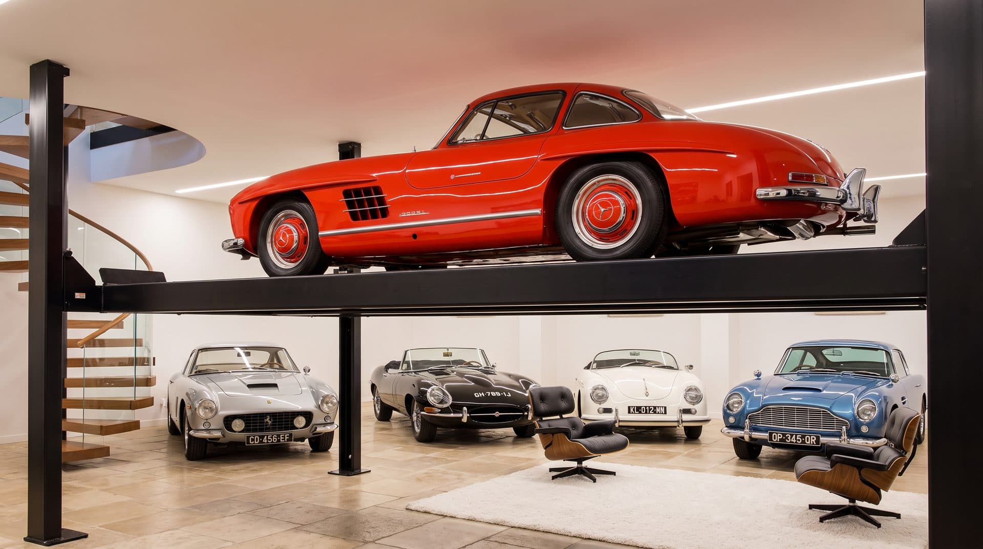 Galerie de collection privée : coupé rouge ancien levé sur un plateau d'exposition, quatre voitures de collection rangées dessous