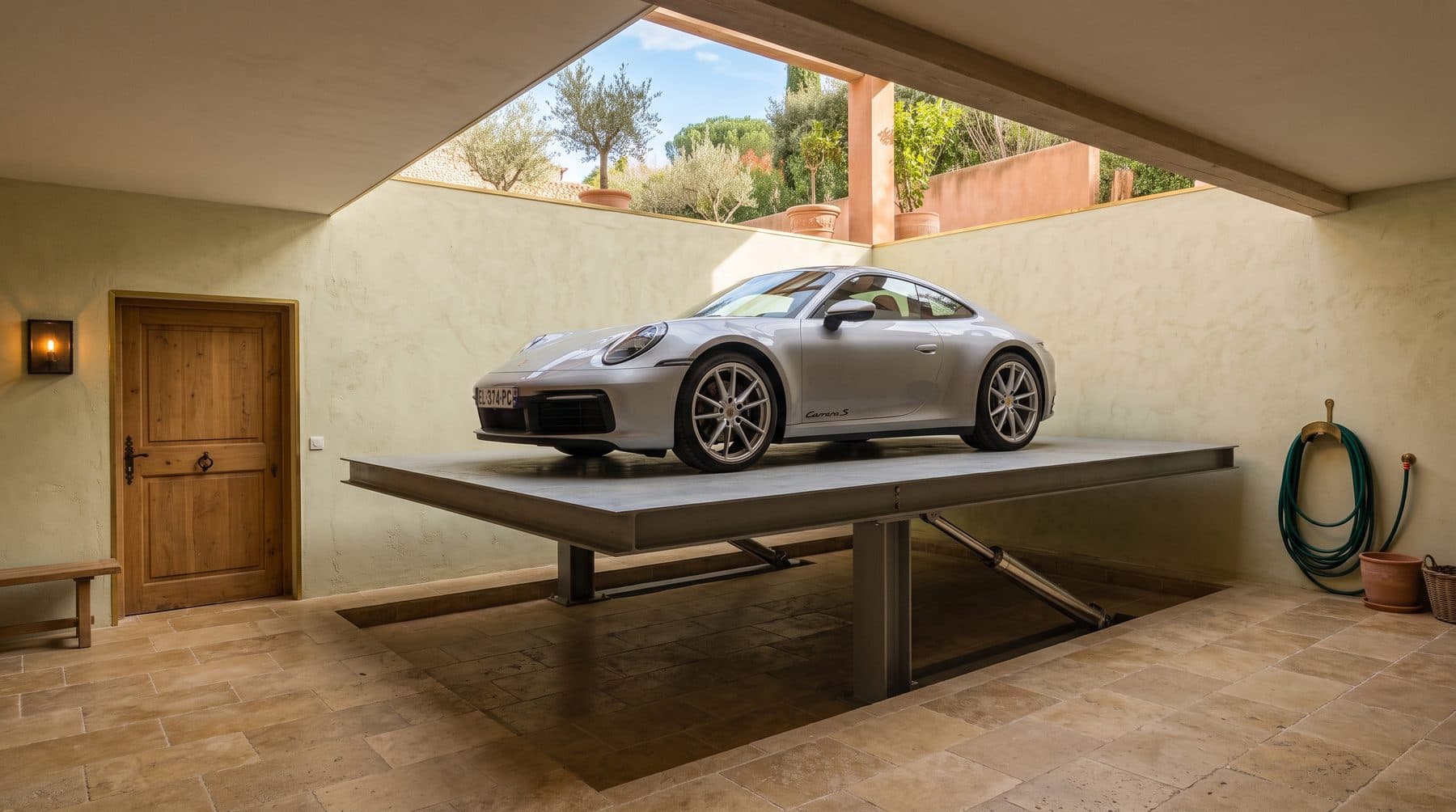 Coupé de sport sur la plateforme d'un monte-voiture dans le garage enterré d'une villa méditerranéenne, trémie ouverte sur le jardin et ses oliviers