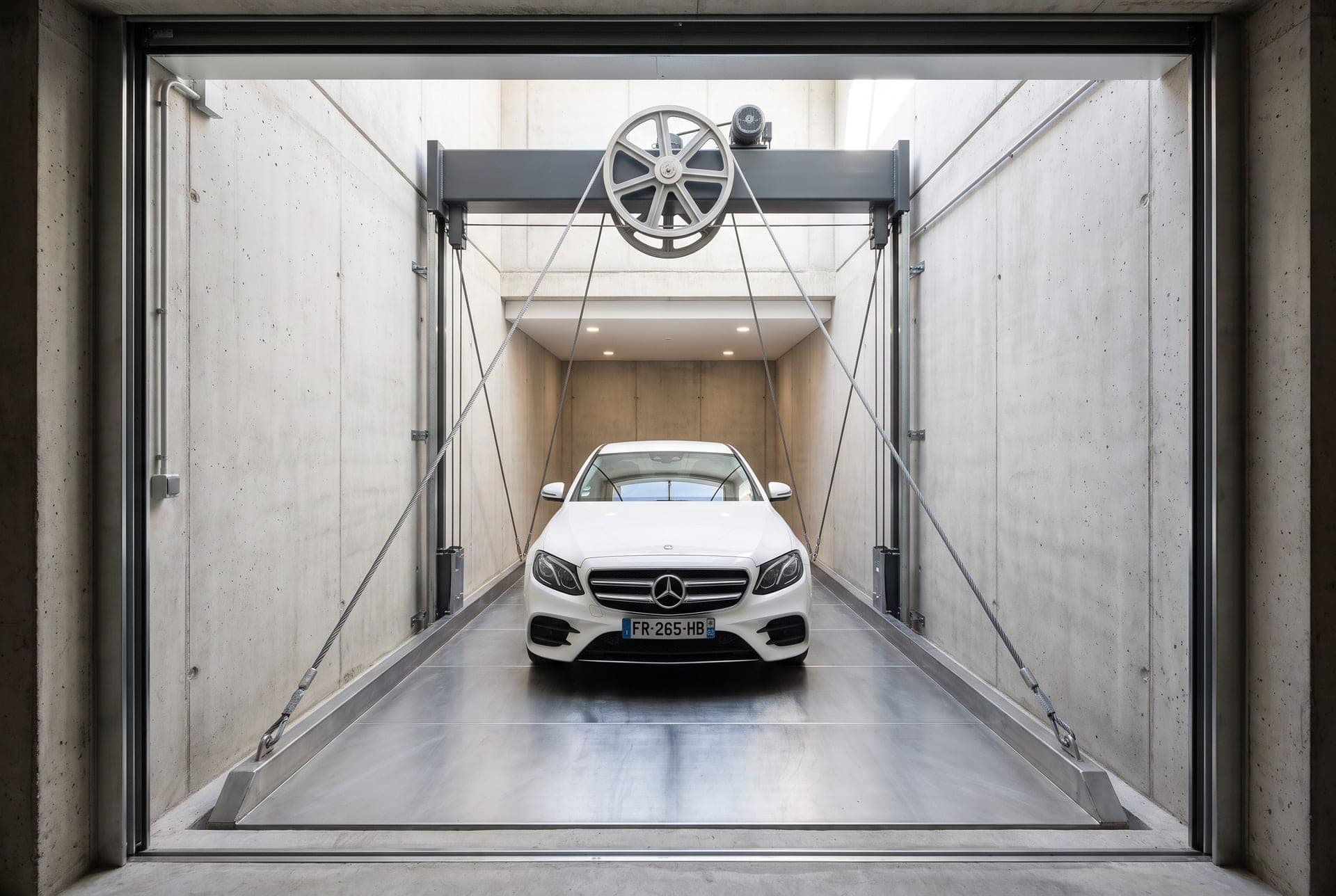Monte-voiture à câbles entier : berline blanche sur la plateforme au pied de la gaine, câbles acier remontant vers la poulie et la poutre de tête